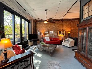 Ein geräumiges Wohnzimmer mit Backsteinwand, großen Fenstern, Deckenventilator und Kamin.
