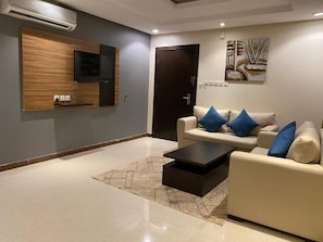 A modern living room with a flat-screen TV mounted on a wooden panel, a beige sofa with blue cushions, a black coffee table, and a patterned rug. - Alrafahya Crown (Riyadh)
