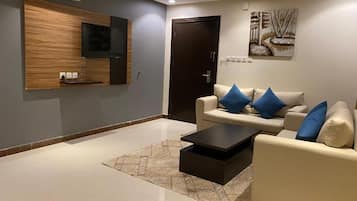 A modern living room with a flat-screen TV mounted on a wooden panel, a beige sofa with blue cushions, a black coffee table, and a patterned rug.