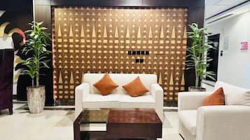 A modern lobby with a patterned wall, two potted plants, a white sofa with orange cushions, and a glass coffee table.