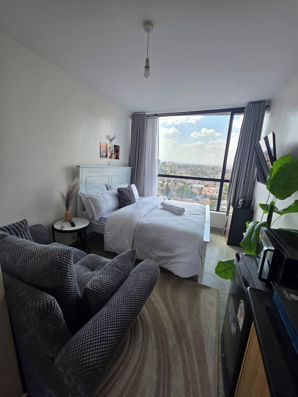 A hotel room with a bed, a sofa, a small table, and a large window offering a city view.