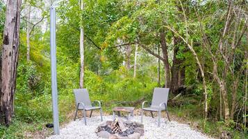 An outdoor seating area with two chairs and a small table, surrounded by trees and a fire pit.