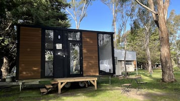 A modern, compact mobile home with a black exterior and wooden accents, situated in a grassy area with trees in the background.