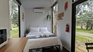 A bedroom with a bed, a wooden table, a microwave, a fire extinguisher, and a view of the outdoors.