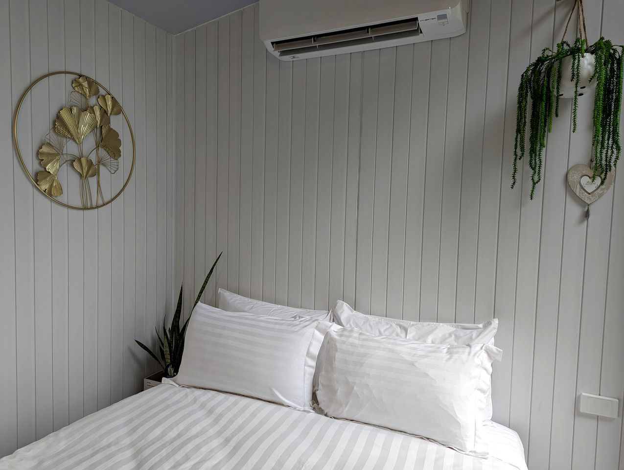 A bedroom with a bed, a wall-mounted air conditioner, a decorative wall piece, and a hanging plant.
