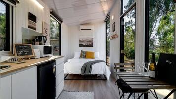 A compact room with a bed, a small table, a chair, a kitchen area, and a view of the outdoors.