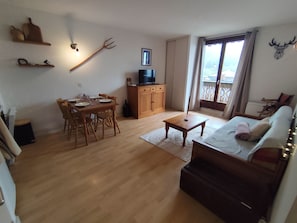 Ein Wohnzimmer mit Holzboden, einem Esstisch mit Stühlen, einer Couch, einem Fernseher auf einem Holzschrank und einem Fenster mit Vorhängen.
