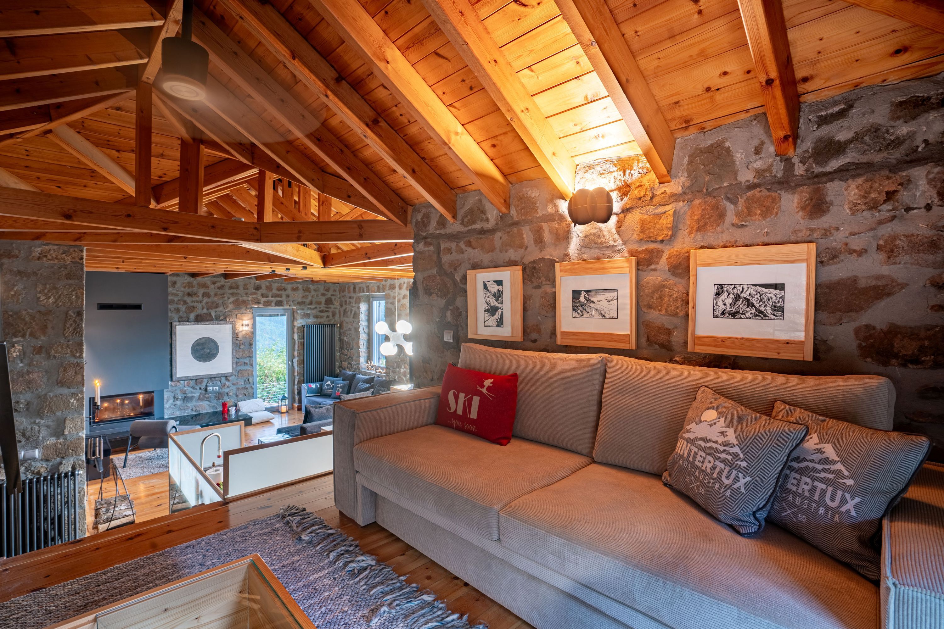 Ein gemütliches Wohnzimmer mit Steinwand, Holzbalken, einer Couch mit Kissen und gerahmten Bildern.