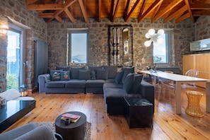 A cozy living room with a stone wall, wooden ceiling beams, a sectional sofa, a dining table with chairs, and a view of the outdoors.