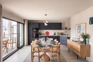 A modern kitchen with dark cabinets, a dining area with a round table and chairs, and a view of the outdoors through large sliding doors. - L'Écrin du Homard Bleu - Plein centre de Dinard (Dinard)