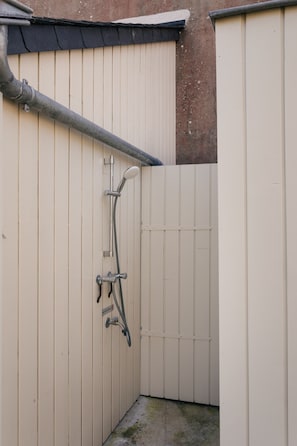 Uma área de duche exterior pequena e fechada, com um chuveiro de metal, um acessório de duche manual e uma torneira.