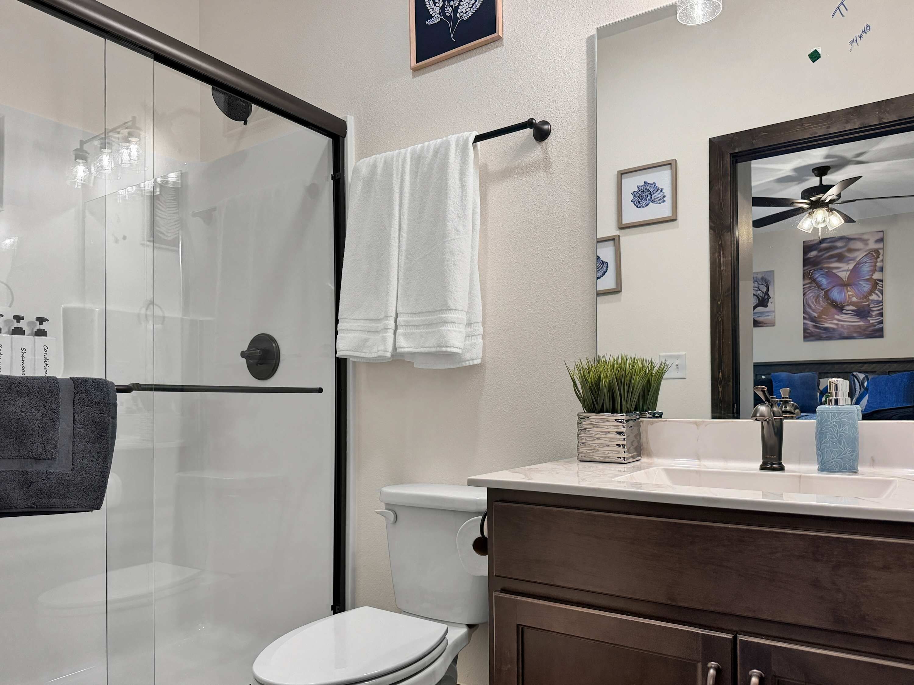 Une salle de bain avec une douche à parois de verre, des toilettes, un meuble-lavabo avec un miroir et un ventilateur de plafond.