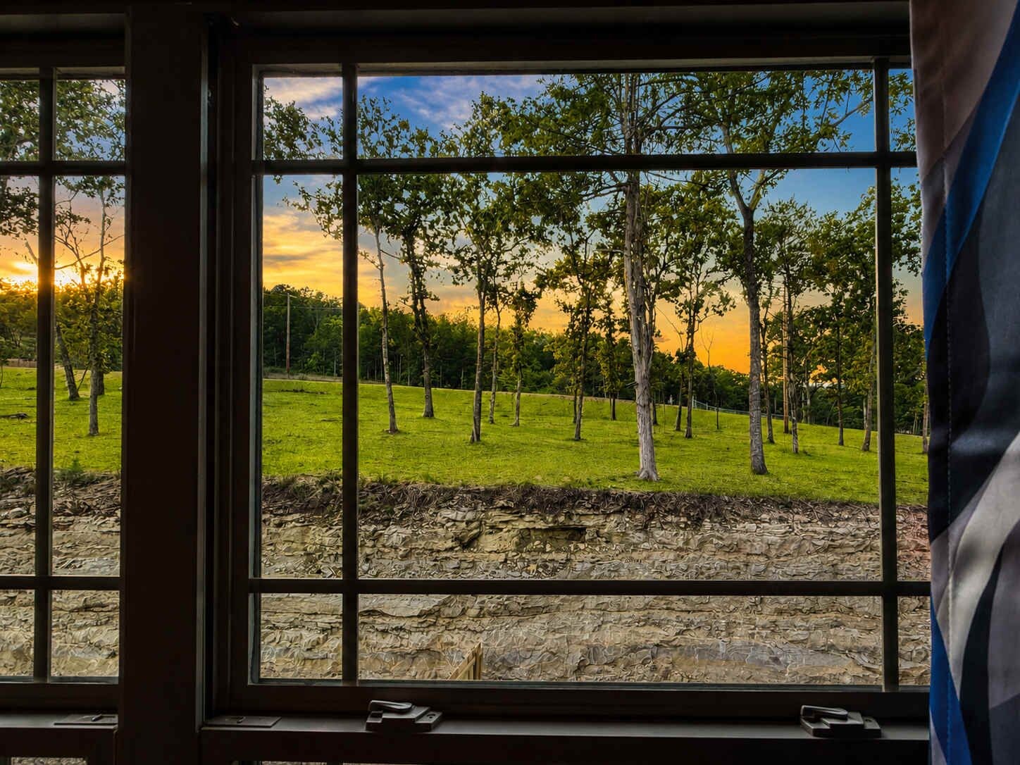 A window with a view of a grassy field and trees, with a stone wall at the bottom.