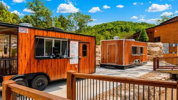 Un camion-restaurant en bois, aménagé comme un véhicule récréatif, avec un panneau affichant le menu, est stationné dans un espace paysager agrémenté d’une clôture en bois et d’un bâtiment en arrière-plan.