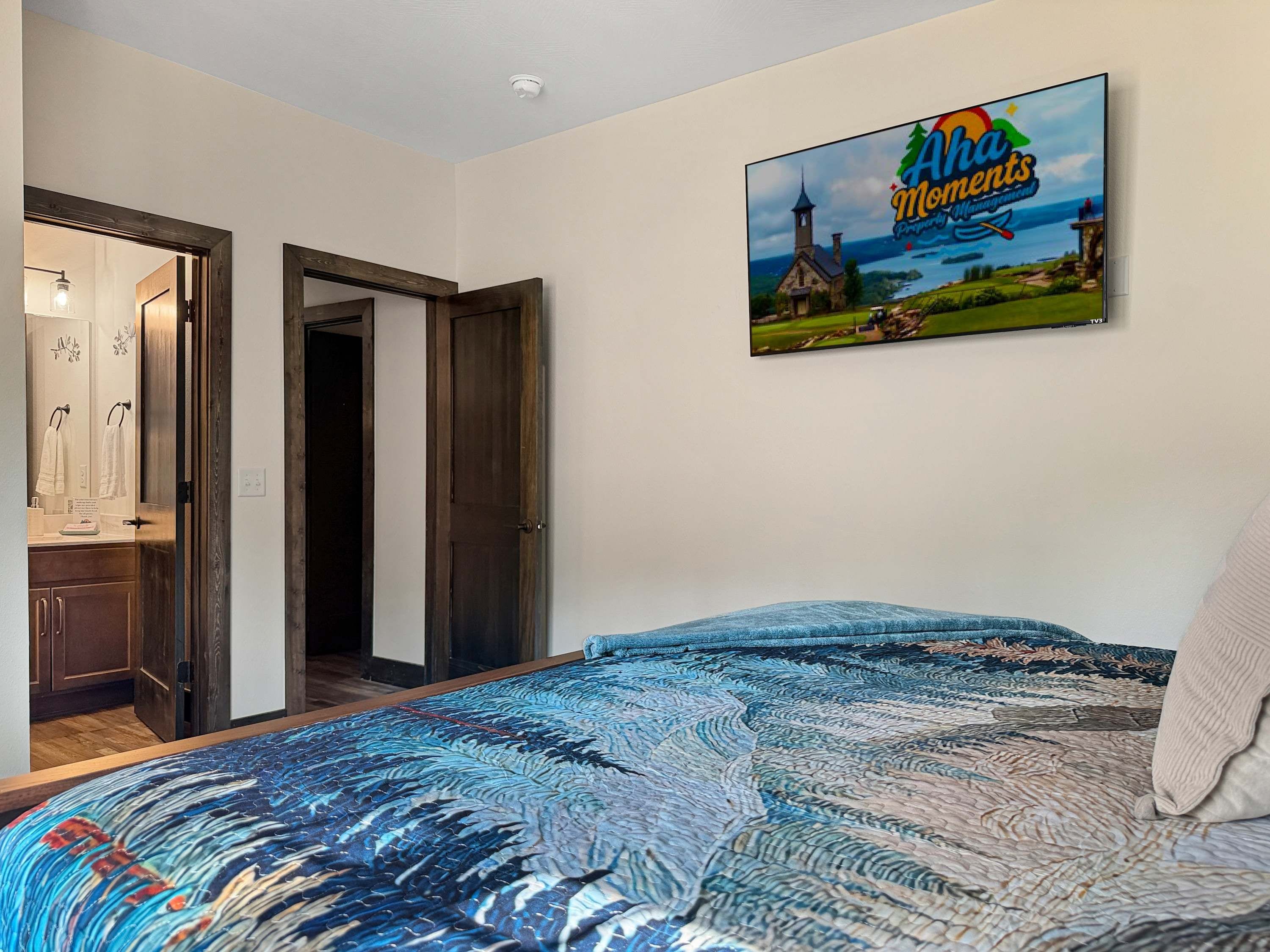 Ein Schlafzimmer mit einem Bett, einem an der Wand befestigten Fernseher und einer Tür zu einem anderen Raum.