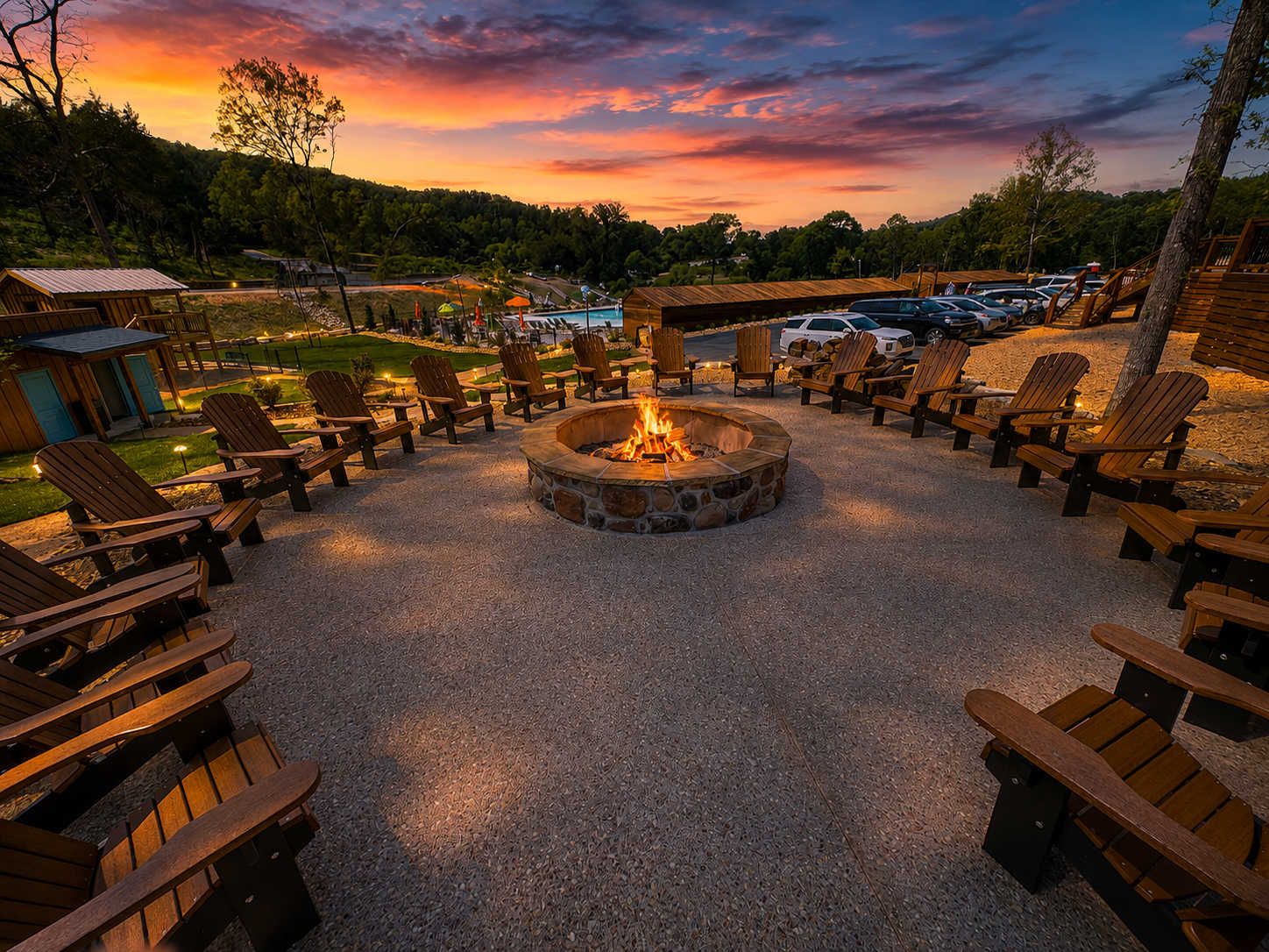A fire pit surrounded by Adirondack chairs, a gravel area, and a parking lot with cars.