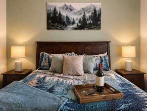 Een slaapkamer met een schilderij van een berglandschap, een houten hoofdbord, twee bedlampen, een bed met een geborduurde spreidekker en een dienblad met een wijnfles en glazen.
