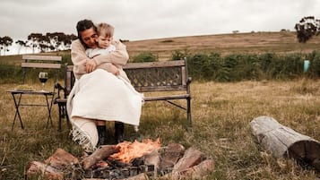 A person sitting on a bench with a child, a fire pit in the foreground, and a grassy field in the background.