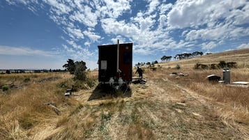 Una cabaña aislada con un cobertizo pequeño, rodeada de pastizales secos y pocos árboles bajo un cielo parcialmente nublado.