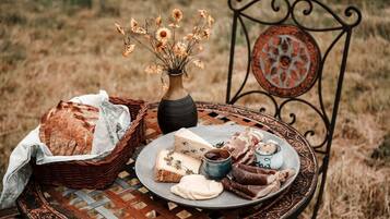 A rustic outdoor setting with a wicker basket, a plate of cheese, a small bowl of jam, and a vase with dried flowers on a metal table.