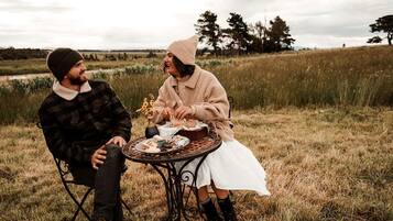 Two people sitting at a round table outdoors with a grassy field and trees in the background.