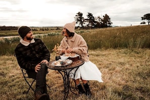 Two people sitting at a round table outdoors with a grassy field and trees in the background.