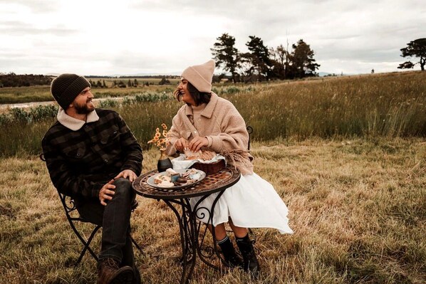 Deux personnes sont assises à une table ronde à l’extérieur, avec un champ herbeux et des arbres en arrière-plan.