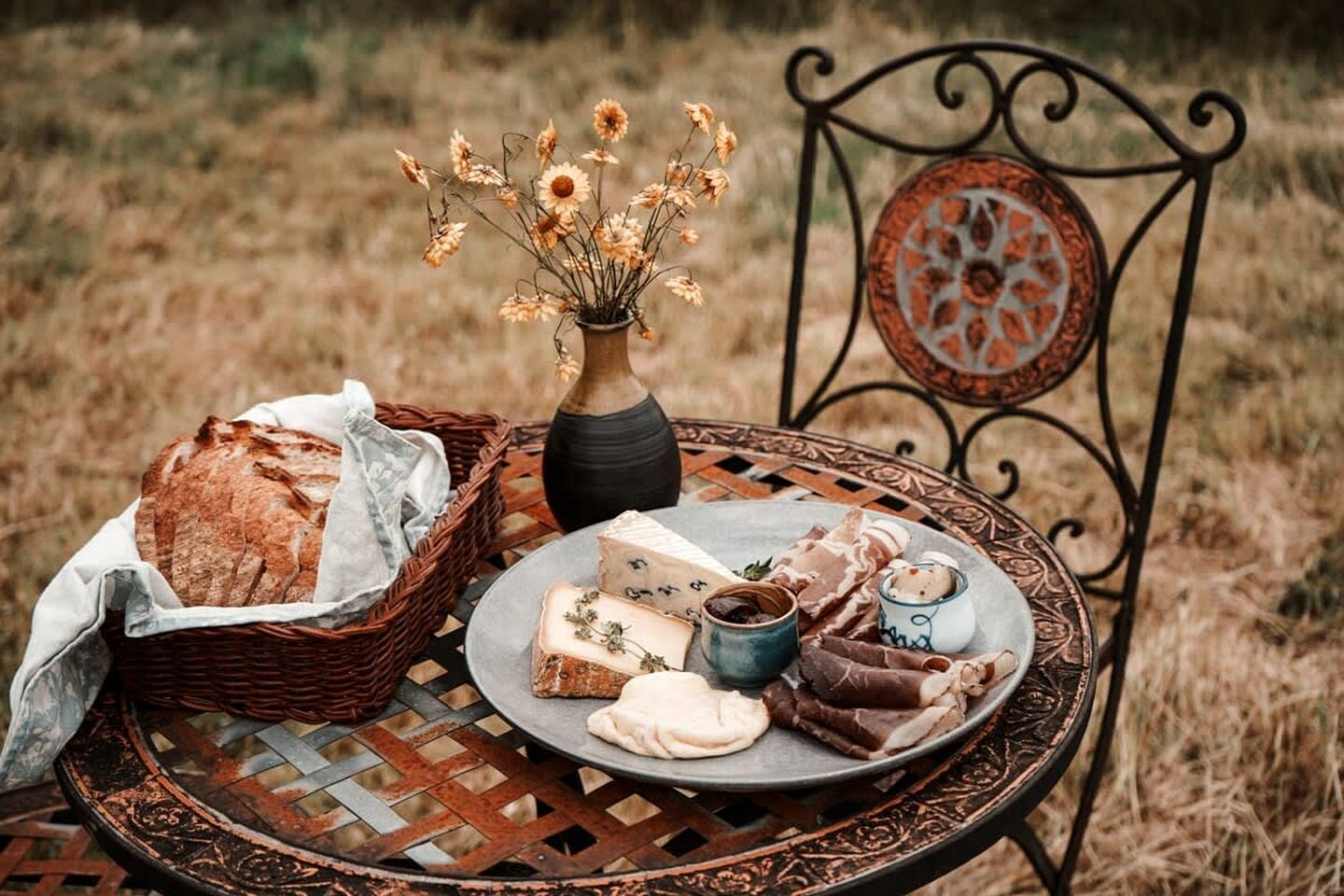 A rustic outdoor setting with a wicker basket, a plate of cheese, a small bowl of jam, and a vase with dried flowers on a metal table.