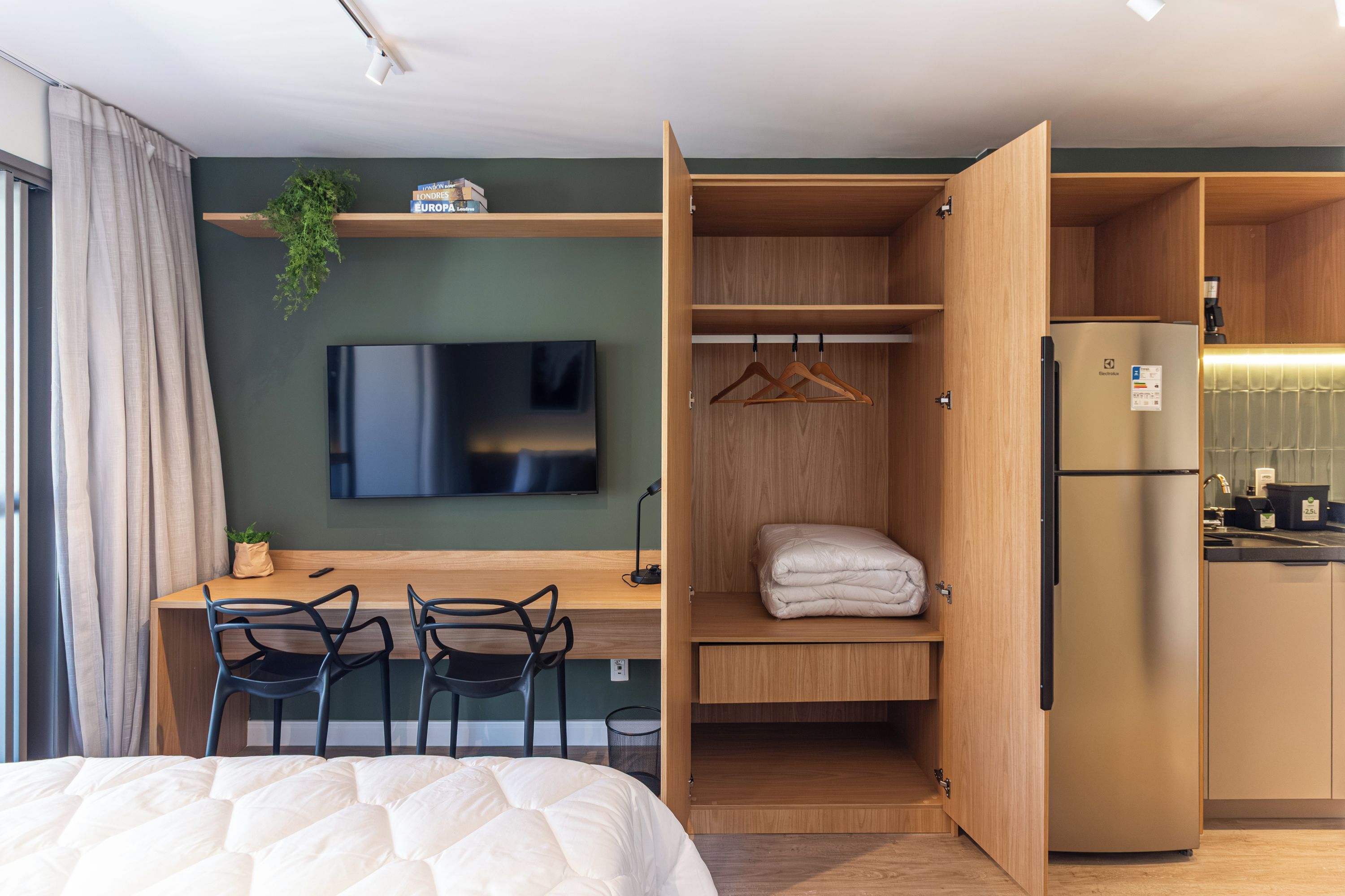 Quarto de hotel compacto, com cama, escrivaninha com cadeiras, televisão, cozinha compacta e guarda-roupa.