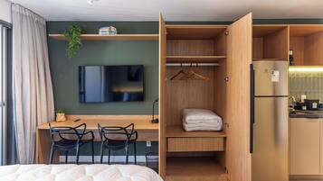 Quarto de hotel compacto, com cama, escrivaninha com cadeiras, televisão, cozinha compacta e guarda-roupa.