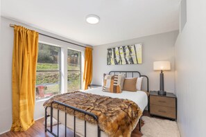A bedroom with a bed, a nightstand, a lamp, a painting, and a window with curtains.