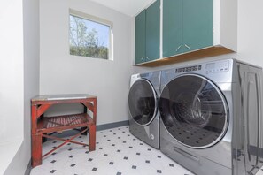 A laundry room with two front-loading washers and dryers, a small wooden shelf, and a window with a view of trees.
