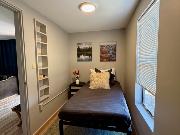 Un dormitorio con cama, mesita de noche con un florero, dos cuadros en la pared y una ventana con persianas.