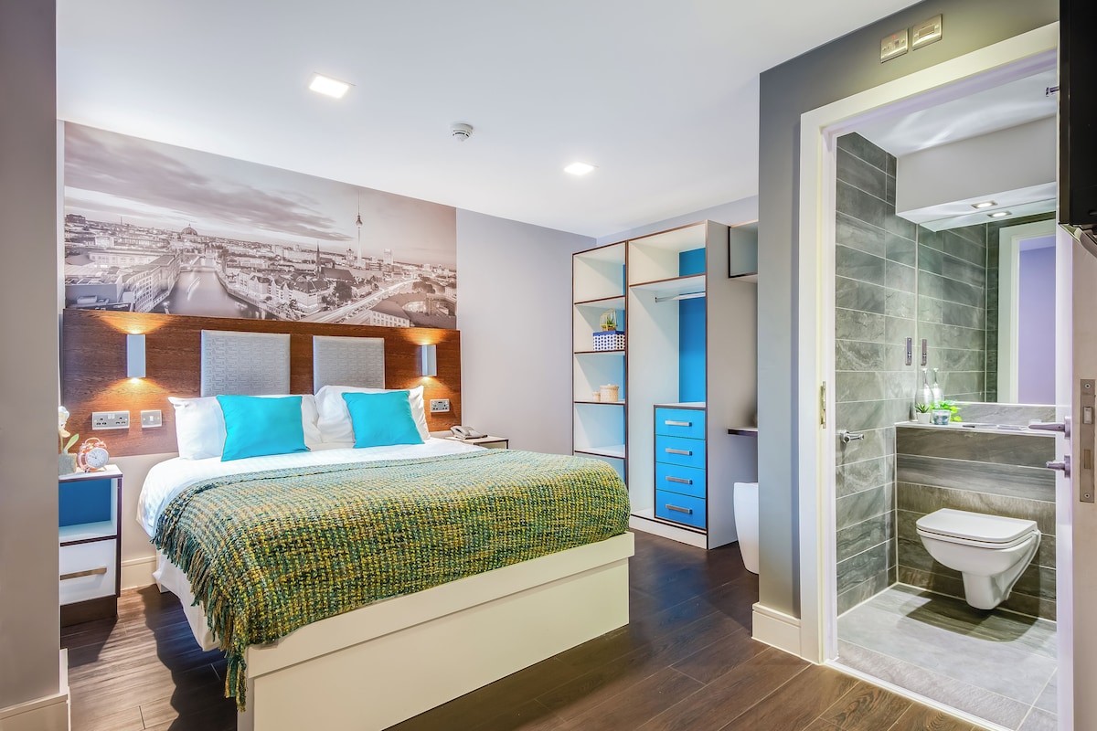 Quarto de hotel moderno com cama grande, painel artístico com vista urbana, closet espaçoso e banheiro separado.