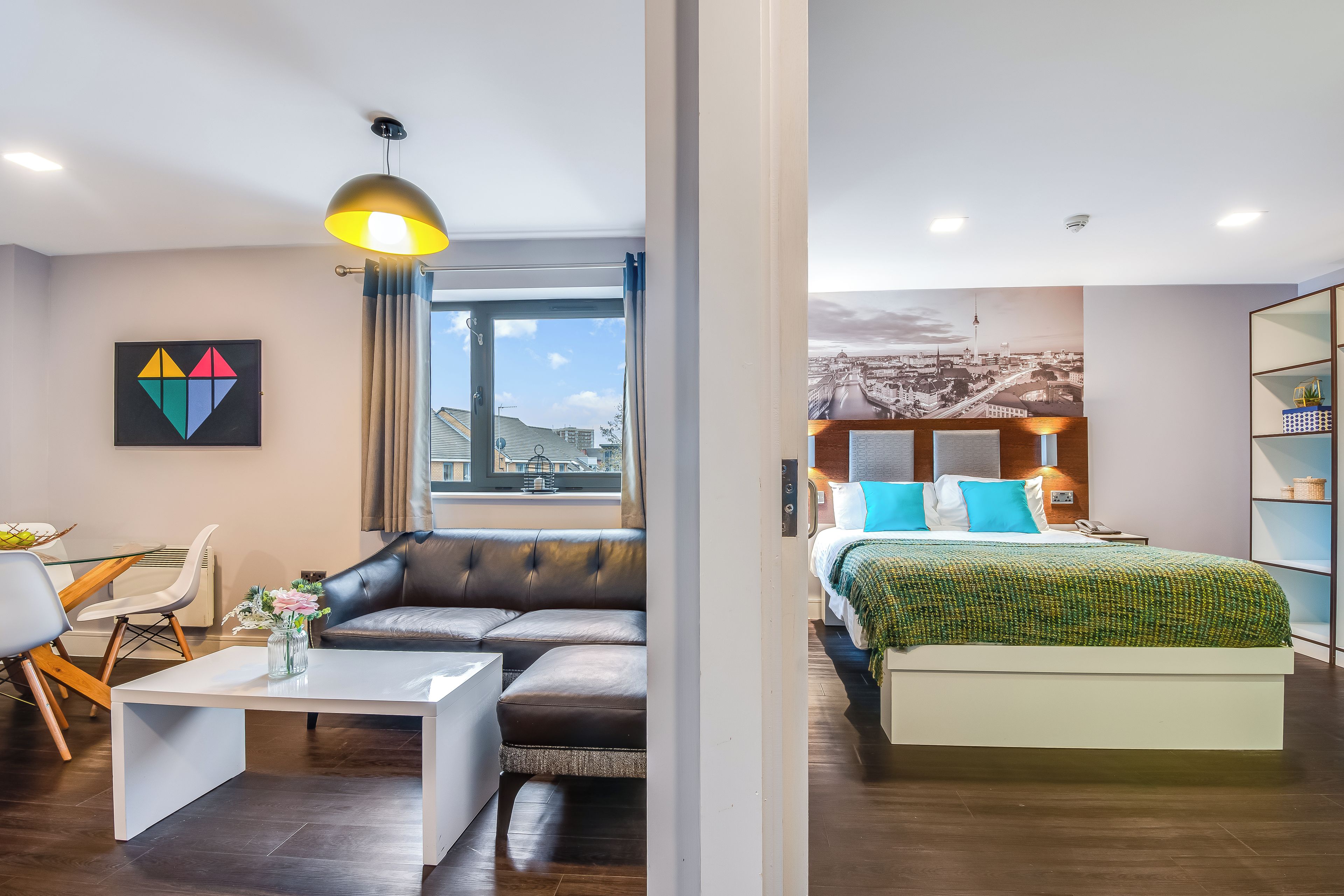 Habitación de hotel moderna con sofá, mesa de comedor, cama con manta verde y vista a la ciudad.