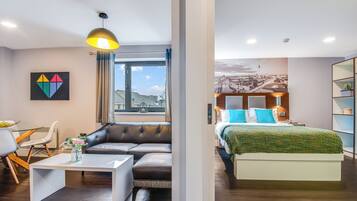 Habitación de hotel moderna con sofá, mesa de comedor, cama con manta verde y vista a la ciudad.