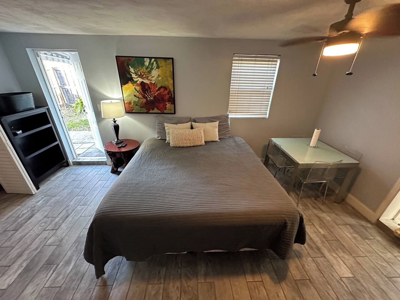 Quarto com cama, escrivaninha, cadeira, abajur, quadro e janela com persianas.