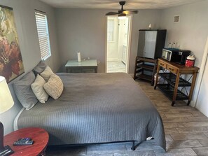 Un dormitorio con cama, mesita de noche, lámpara y una pequeña mesa de madera con microondas y cafetera.