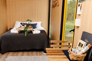 Ein modernes Schlafzimmer mit einem großen Bett, Holznachtschränkchen, einem kleinen Holztisch mit Handtüchern und einer Pflanze sowie Blick ins Freie durch eine Glastür.