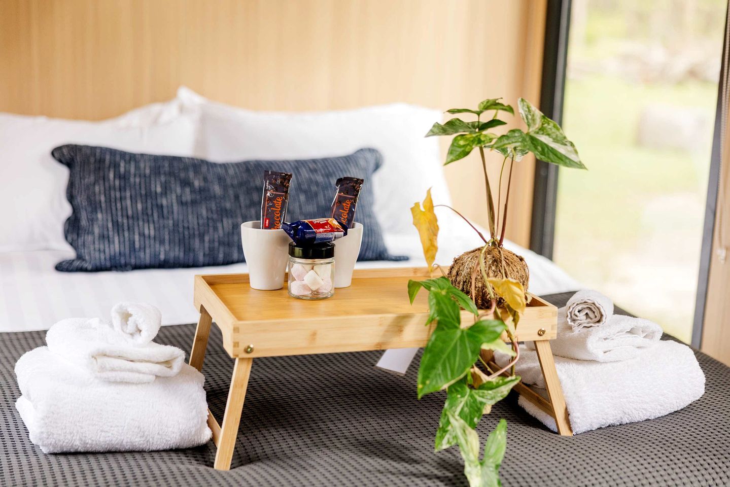A neatly made bed with white linens, a tray with chocolate bars, a small glass jar, and a potted plant.