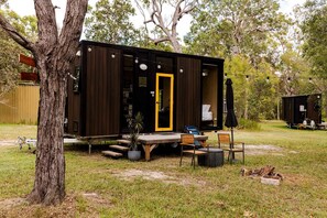A small, dark wooden cabin with a yellow door, a picnic table, and a few chairs outside. - Cozy Tiny House Retreat with Firepit & Bush Views in Round Hill, Australia (Round Hill)