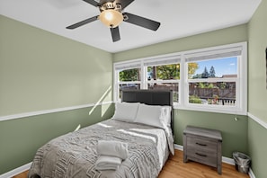 Une chambre à coucher avec un lit, une table de chevet, un ventilateur de plafond et une fenêtre donnant sur l’extérieur.