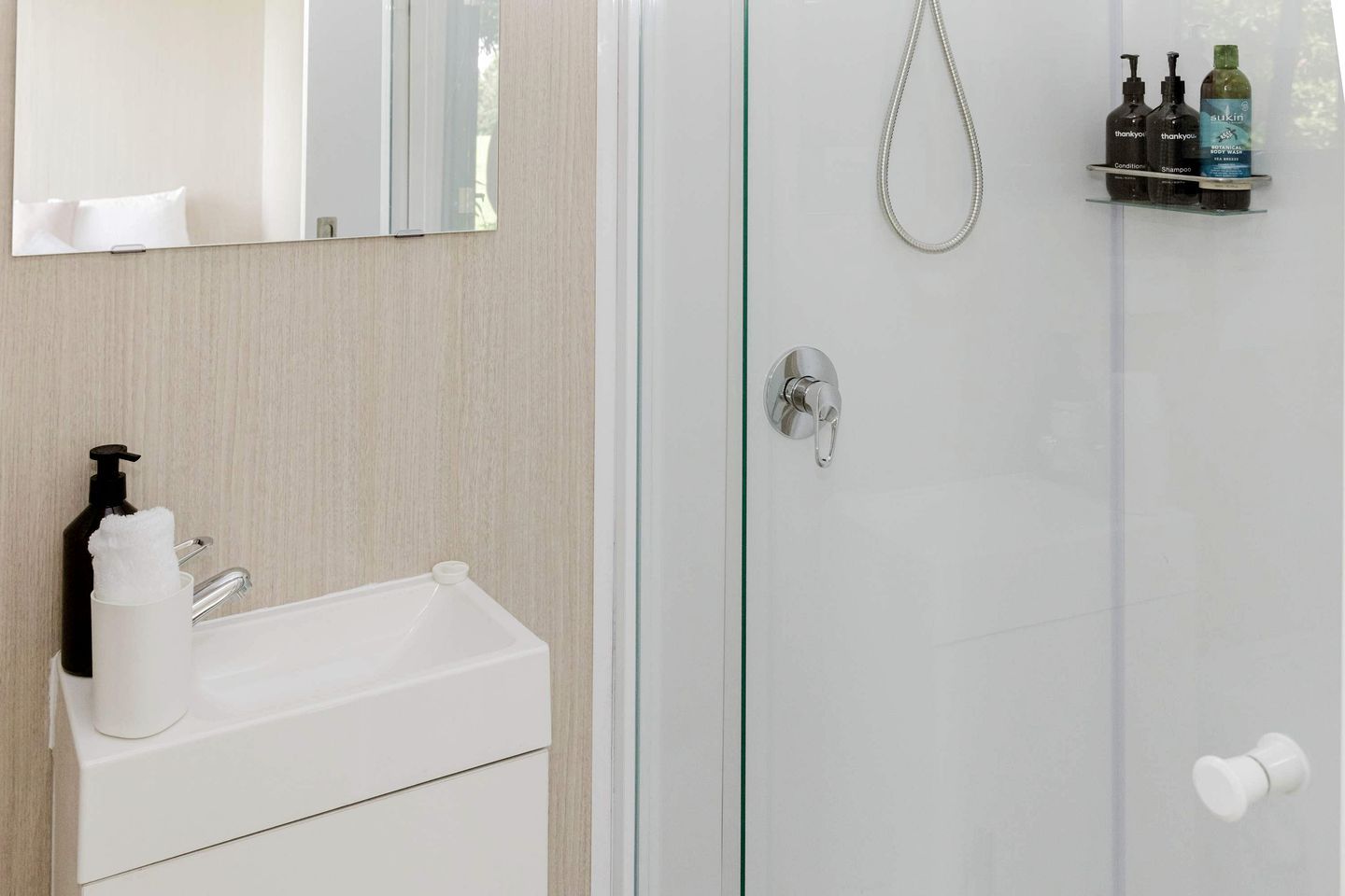 Et badeværelse med en hvid vask, et spejl, en bruseniche med glasdør og toiletartikler på en hylde.