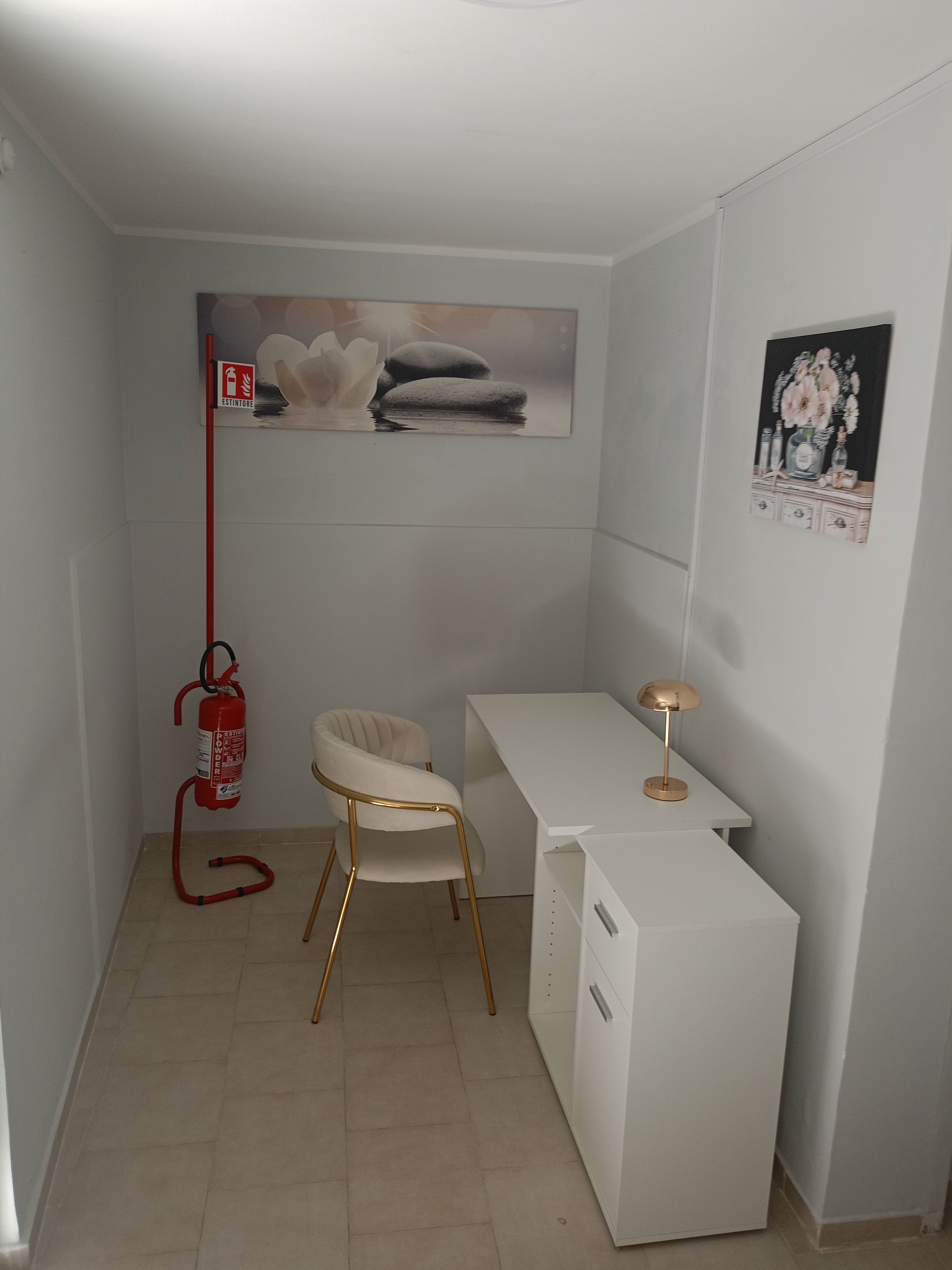 Une petite pièce blanche, avec un bureau, une chaise, un extincteur et un tableau accroché au mur.