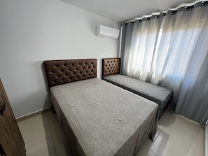 Quarto com duas camas, ar condicionado na parede e janela com cortinas.