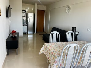 A modern living room with a dining table, a sofa, a TV mounted on the wall, and a refrigerator in the background. - CRCN - Amethist Tower (Caldas Novas)