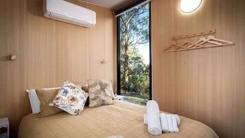 Une chambre à coucher comprenant un lit, des oreillers, une table de chevet, un appareil de climatisation, une fenêtre donnant sur des arbres et un luminaire fixé au mur.