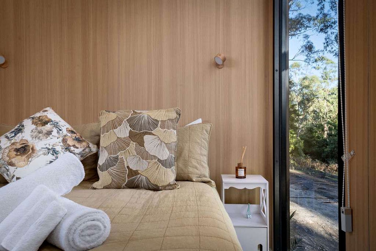 Une chambre à coucher avec un lit, des oreillers, une table de chevet avec une bouteille, et une vue sur l’extérieur.