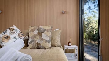 Une chambre à coucher avec un lit, des oreillers, une table de chevet avec une bouteille, et une vue sur l’extérieur.
