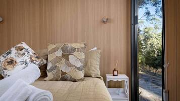 A bedroom with a bed, pillows, a bedside table with a bottle, and a view of the outdoors.